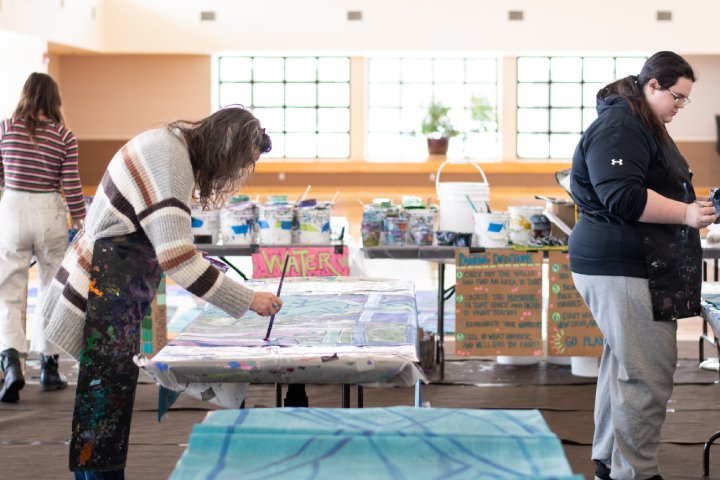 People painting large canvases in a spacious, well-lit room. Supplies, including paint buckets and brushes, are spread across tables, and signs with instructions are visible. The participants are focused on their mural work, wearing aprons to protect their clothes.